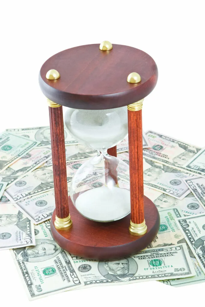 Time is money illustrated by an hourglass sitting on top of a stack of $50 bills. 