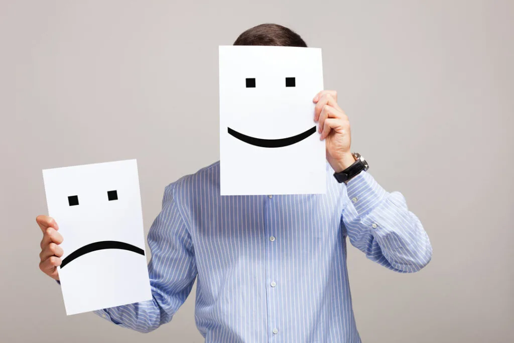 Person holding up a smiley face over his face, and a frown face paper held in the other hand.