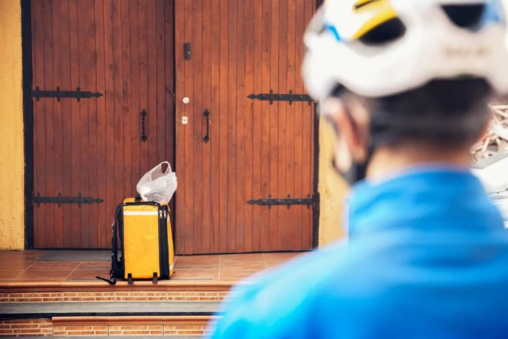 A courier in a bike helmet stands back from the door after leaving a contactless delivery at the customer's door.