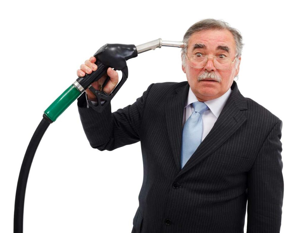 A man in a business suit holding a gas nozzle up to his head like he's ready to shoot himself with it.