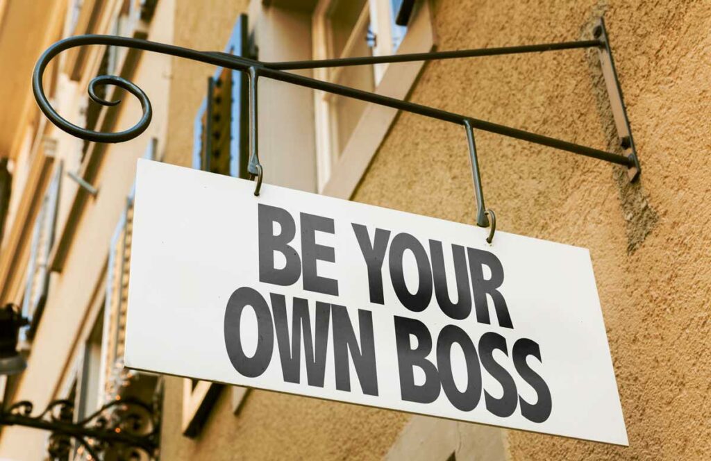 An old fashioned hanging business sign hangs against an older stucco office building. The sign reads Be your own boss.