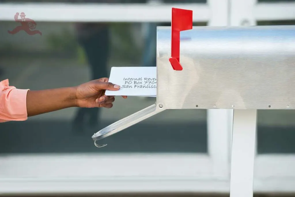 A person's hand putting letters in a mailbox. The top letter includes their estimated tax payment and is addressed to the Internal Revenue Service.