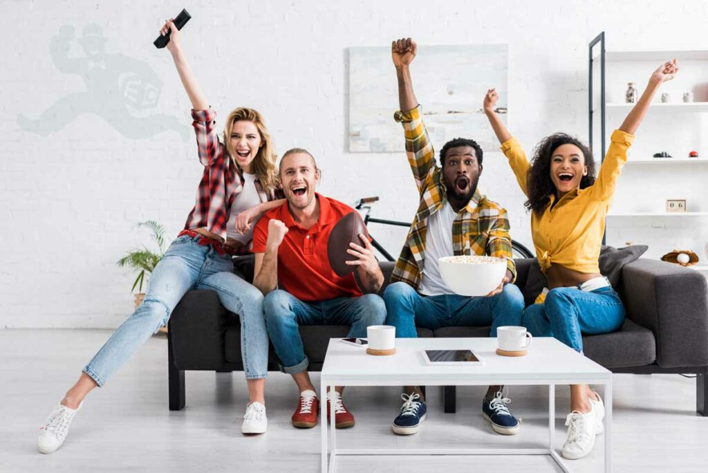 Four friends cheering while watching a football game on TV while thinking of ordering Doordash.