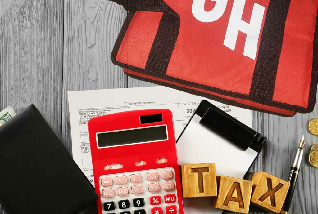 Items showing someone ready to do their Grubhub taxes, with a Grubhub bag, note pad, pen, calculator, billfold, and Grubhub 1099 form all spread out on a table.