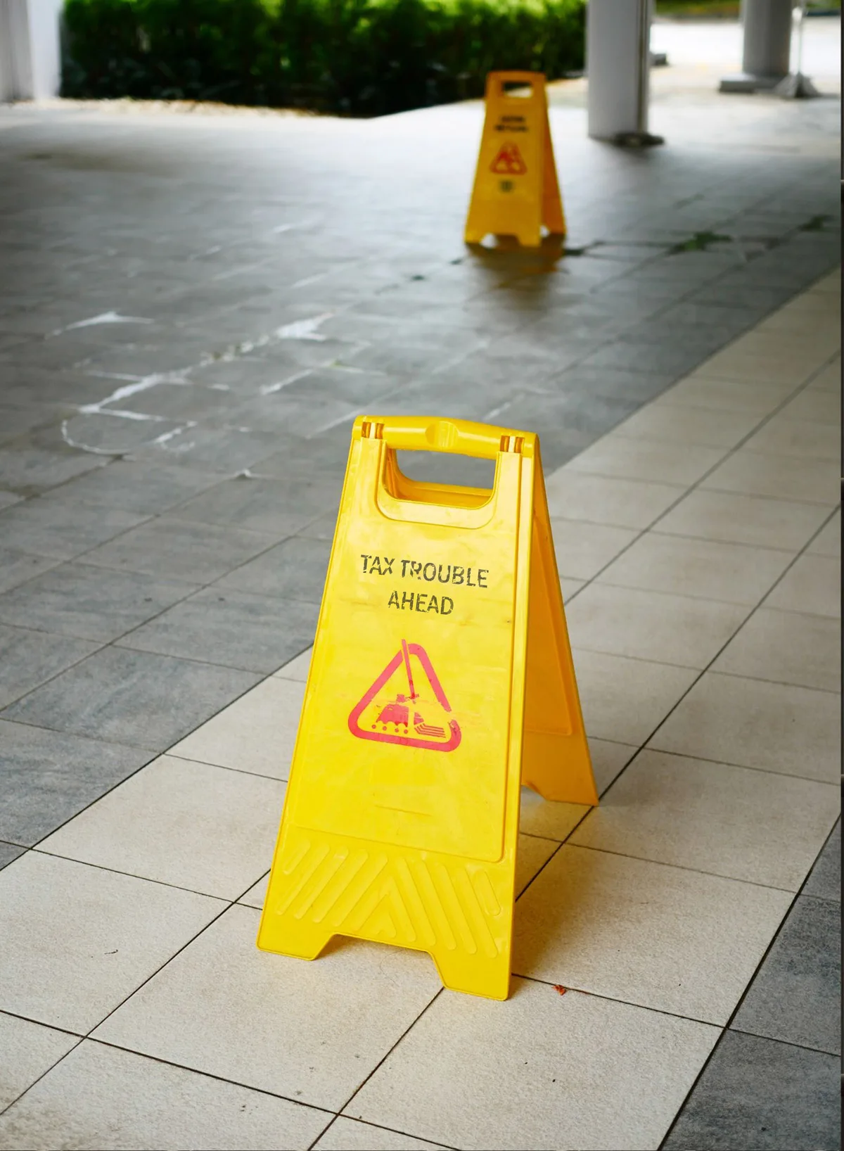 A pair of yellow caution fold up signs on a concrete floor, with the nearest one reading tax trouble ahead.